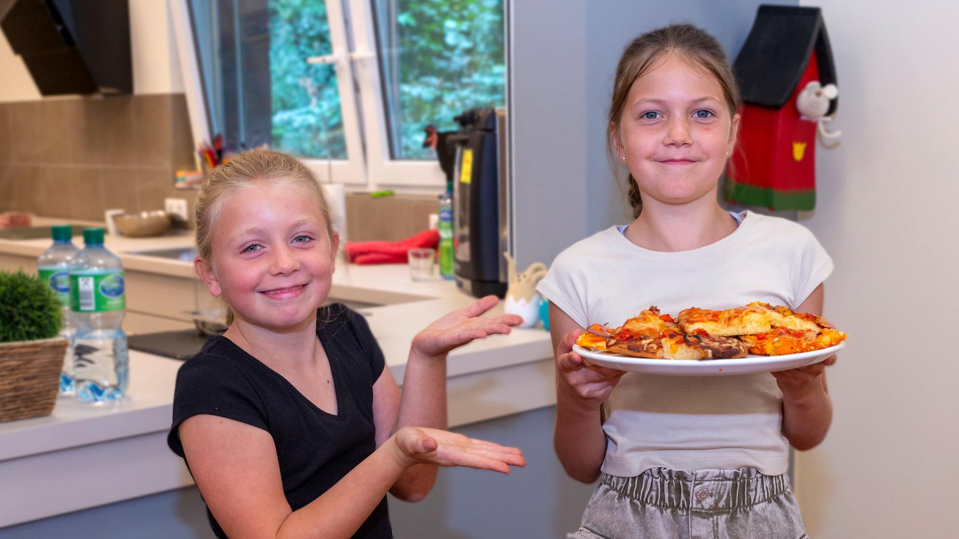 Besseresser Kids - Lege packt´s an: Zwei Schülerinnen stehen in der Schulküche - eine hält präsentierend einen Teller mit Pizza in die Kamera.