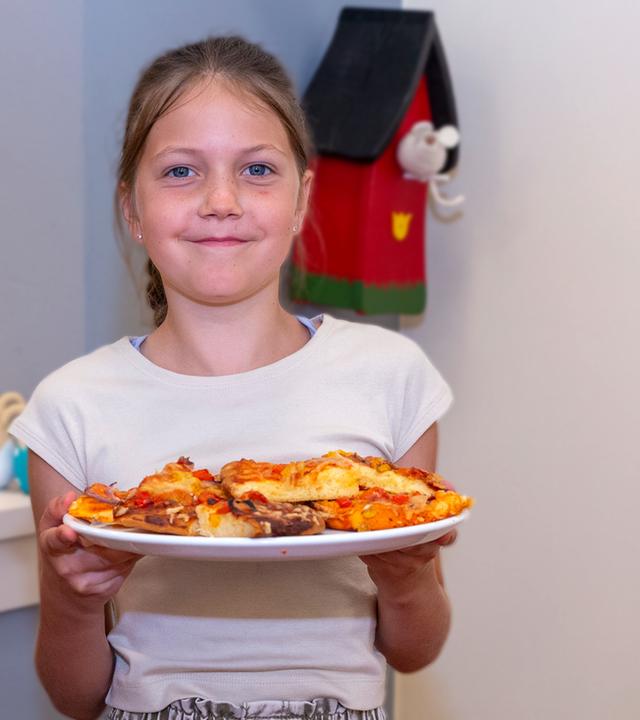 Besseresser Kids - Lege packt´s an: Zwei Schülerinnen stehen in der Schulküche - eine hält präsentierend einen Teller mit Pizza in die Kamera.