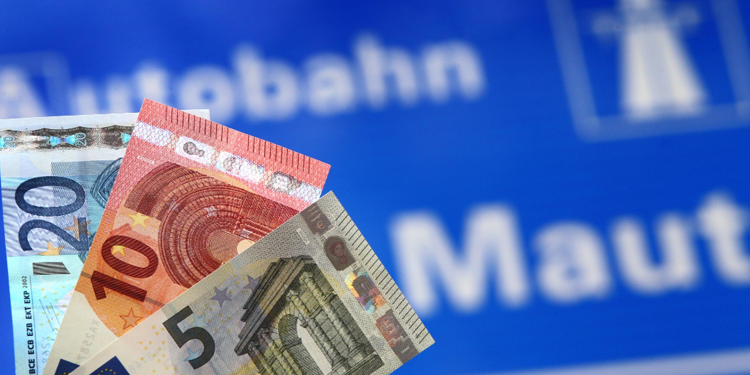Verschiedene Euro-Banknoten werden vor ein Hinweisschild auf Mautpflicht gehalten.