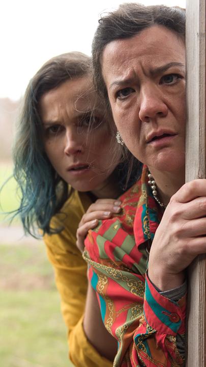 Lin (Karin Hanczewski) und Gloria (Bärbel Schwarz) stehen hinter einer halb geöffneten Tür und blicken neugierig und ungläubig in einen Raum hinein.