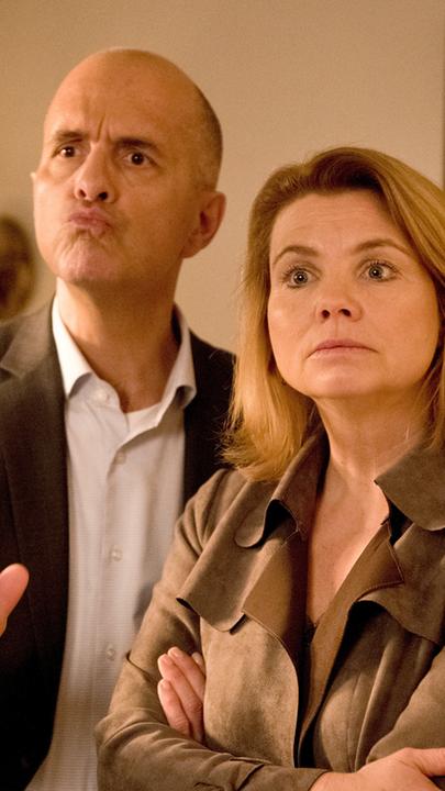 Erik Merz (Christoph Maria Herbst) und Anne Merz (Annette Frier) stehen in einem altmodischen und rustikal eingerichteten Zimmer nebeneinander.