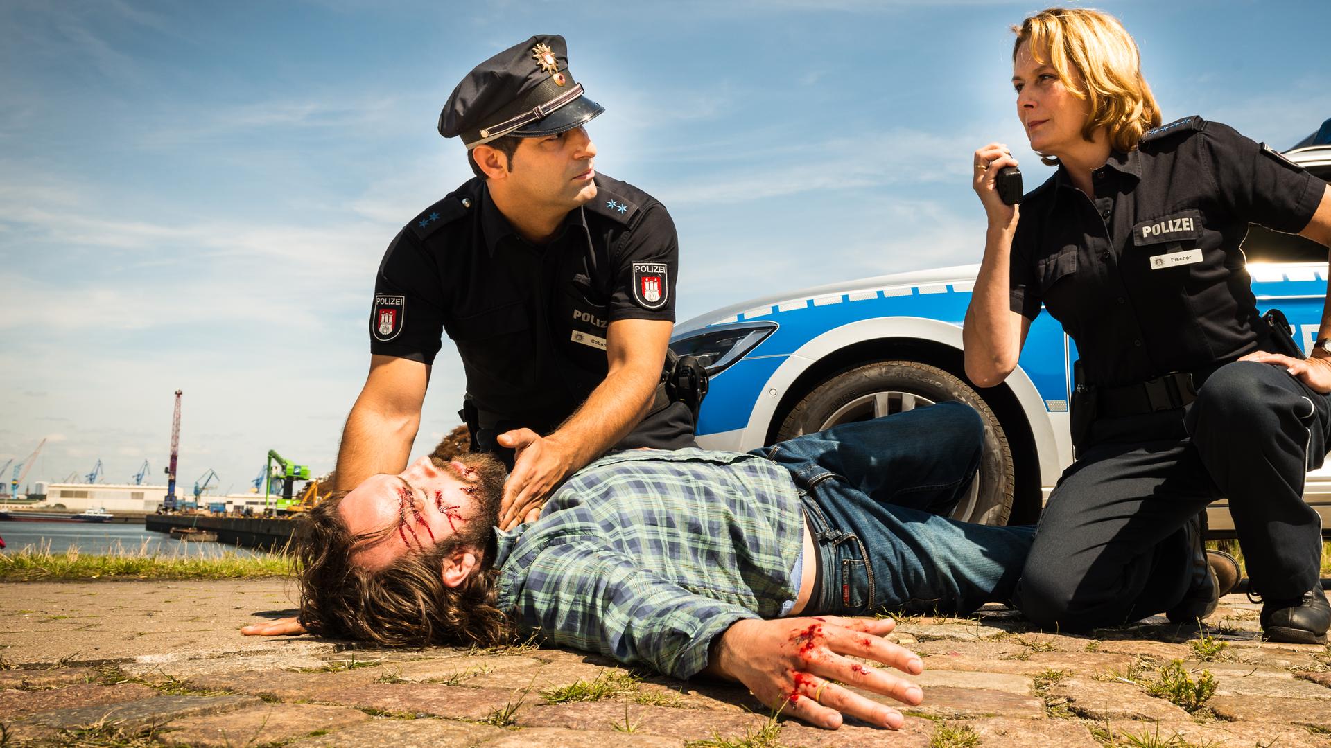 "Notruf Hafenkante - Plan B": Die Polizeibeamten Tarik Coban (Serhat Cokgezen) und Claudia Fischer (Janette Rauch) knien auf dem Boden neben einem auf dem Boden liegenden verletzten Mann (Isaak Dentler). Tarik fühlt den Puls des Mannes und schaut zu seiner Kollegin, die ihr Funkgerät in der Hand hält. Im Hintergrund steht der Streifenwagen.