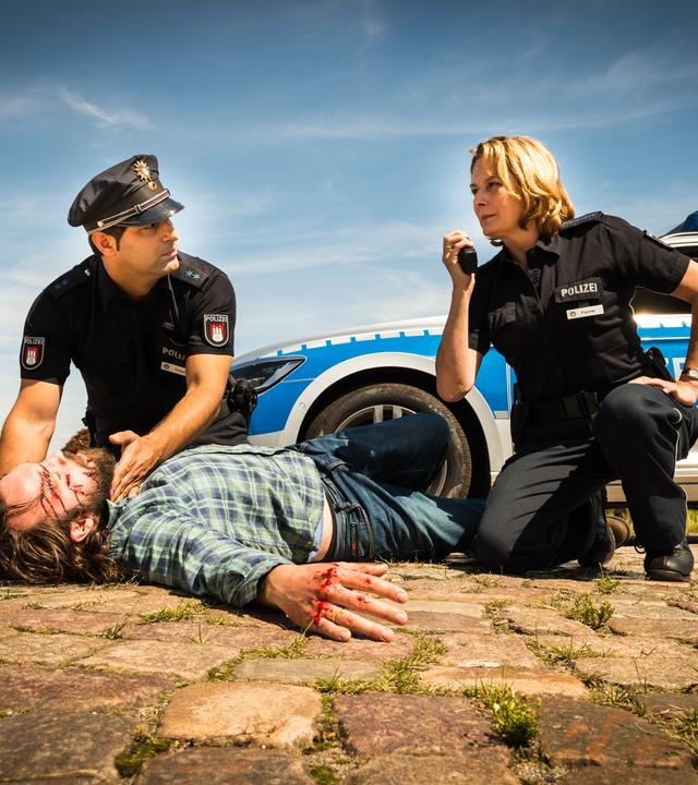 "Notruf Hafenkante - Plan B": Die Polizeibeamten Tarik Coban (Serhat Cokgezen) und Claudia Fischer (Janette Rauch) knien auf dem Boden neben einem auf dem Boden liegenden verletzten Mann (Isaak Dentler). Tarik fühlt den Puls des Mannes und schaut zu seiner Kollegin, die ihr Funkgerät in der Hand hält. Im Hintergrund steht der Streifenwagen.