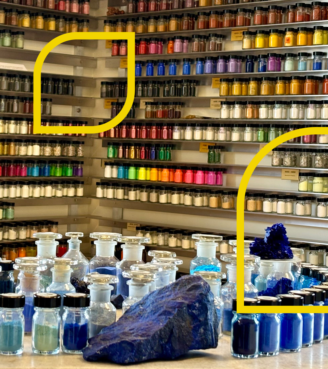 Ein Regal voller Farbgläser. Im Vordergrund stehen auf einem Tisch mehrere Gläser mit Farben in verschiedenen Blautönen.