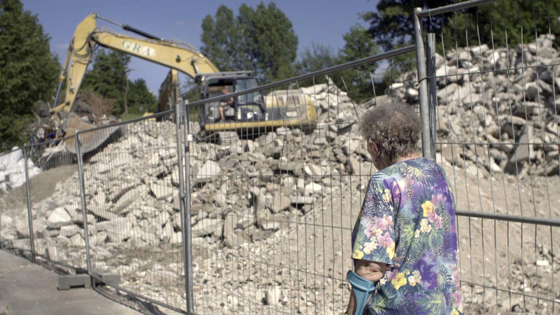 Helga steht vor einem Zaun. Sie trägt eine bunte Bluse und eine Krücke. Hinter dem Zaun ist ein Bagger zu sehen, ein Haus wird abgerissen