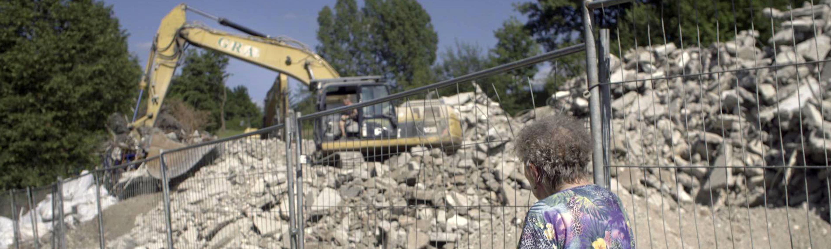 Helga steht vor einem Zaun. Sie trägt eine bunte Bluse und eine Krücke. Hinter dem Zaun ist ein Bagger zu sehen, ein Haus wird abgerissen