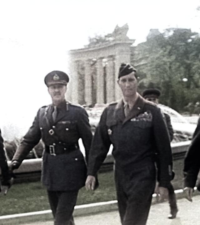 Das Bild zeigt eine Gruppe von vier Soldaten, die in militärischer Uniform gehen. Sie befinden sich auf einem Platz, der von einem Brunnen umgeben ist, und im Hintergrund sind eine Reihe von Säulen oder Gebäuden zu sehen, die auf eine architektonische Struktur hindeuten. Die Soldaten tragen unterschiedliche Uniformen, die auf verschiedene Ränge und Nationalitäten hinweisen könnten. Die Szene wirkt historisch, vermutlich aus der Zeit nach dem Zweiten Weltkrieg. Die allgemeine Atmosphäre vermittelt den Eindruck eines offiziellen Anlasses oder einer Zeremonie. Die Bildqualität ist in Schwarz-Weiß, was den zeitlichen Kontext verstärkt. Es gibt auch einige Bäume und Laternen im Hintergrund, die zur Umgebung beitragen.