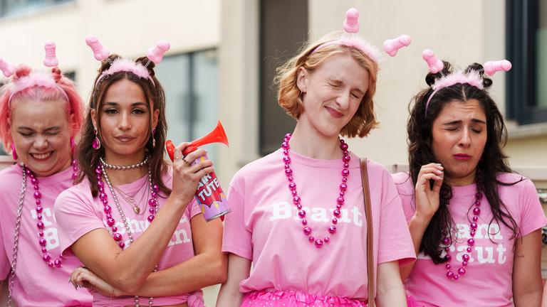 "Plötzlich Schwester": Mira (Jona Pausacker), Sandra (Klara Lange) und Melek (Danai Chatzipetrou) erschrecken sich vor dem Signalhorn, welches Sandy (Cosima Henman) betätigt. Alle vier Frauen stehen vor einem Gebäude und tragen pinke Shirts und Röcke zu Sandras Junggesellinnenabschied.
