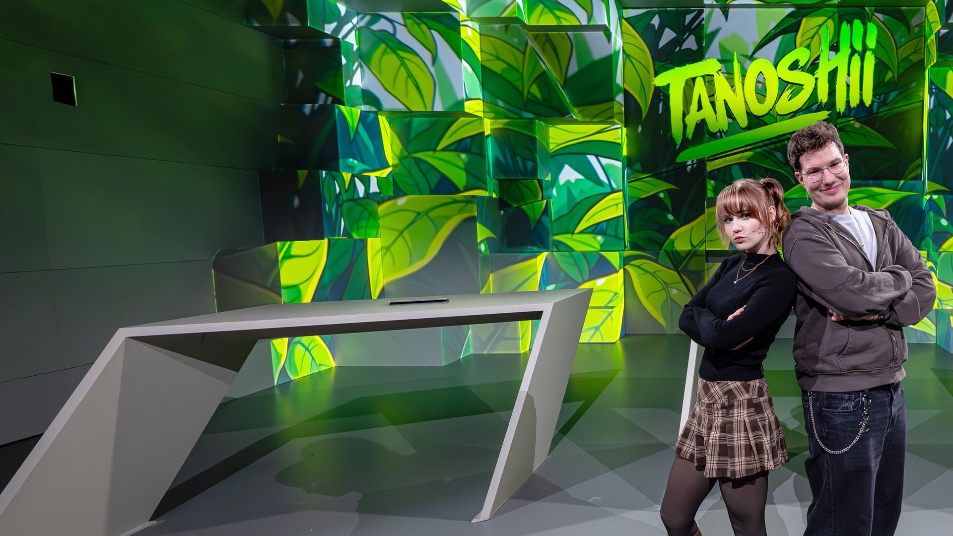 Cosplayerin "Kaddicosplay" (l.) und Creator "TheZeroOfTime" (r.) stehen mit dem Rücken aneinander gelehnt vor der grün gefärbten Studiokulisse. Mittig über ihnen das Tanoshii-Logo.