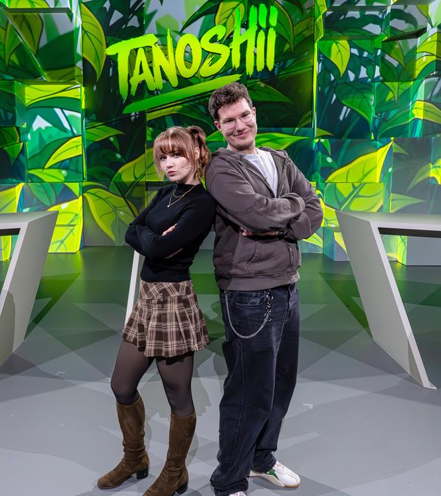 Cosplayerin "Kaddicosplay" (l.) und Creator "TheZeroOfTime" (r.) stehen mit dem Rücken aneinander gelehnt vor der grün gefärbten Studiokulisse. Mittig über ihnen das Tanoshii-Logo.