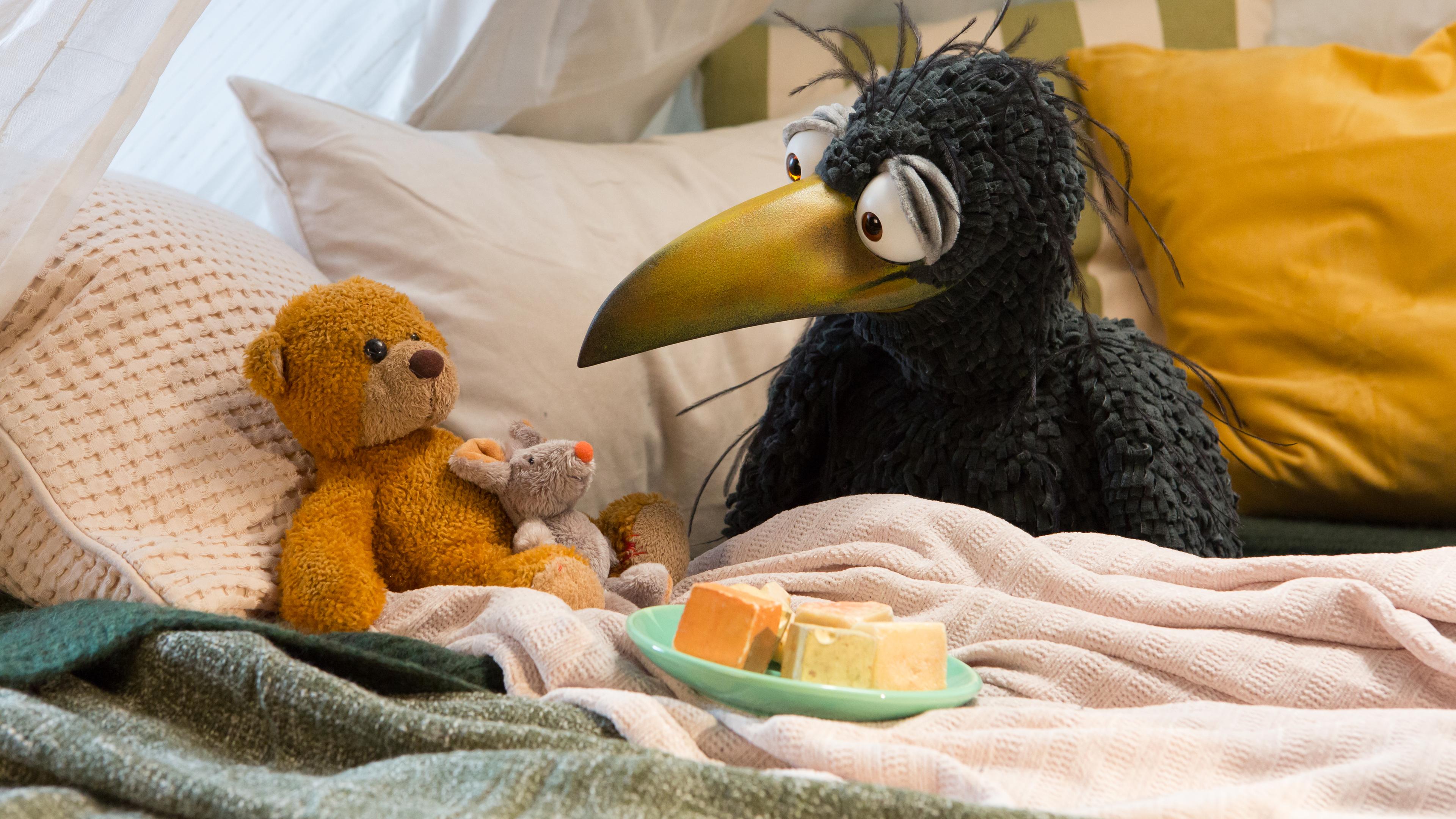 Rabe Rudi und sein Teddy hocken in der gemütlichen Kuschelecke. Vor ihnen steht ein Teller mit Käsehäppchen, auf Teddys Bauch liegt eine Kuschelmaus.