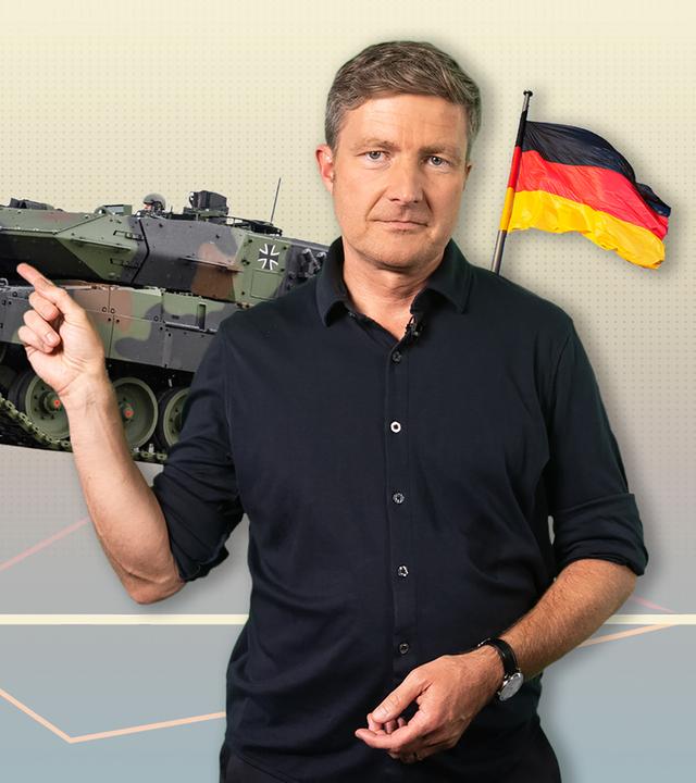 Der Moderator zeigt auf ein Bild eines Panzers in seinem Hintergrund. Rechts ist eine Deutschlandfahne über seiner Schulter abgebildet.