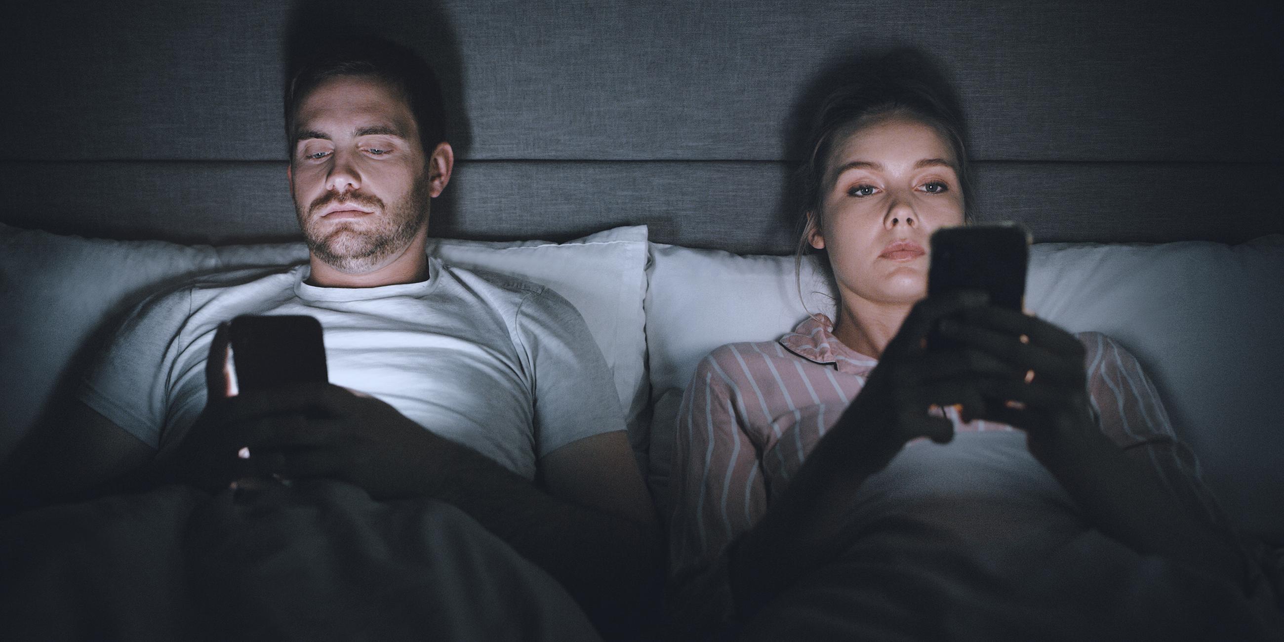 Es sind eine Frau und ein Mann zu sehen. Beide liegen im Bett nebeneinander und schauen intensiv auf ihr Handy.