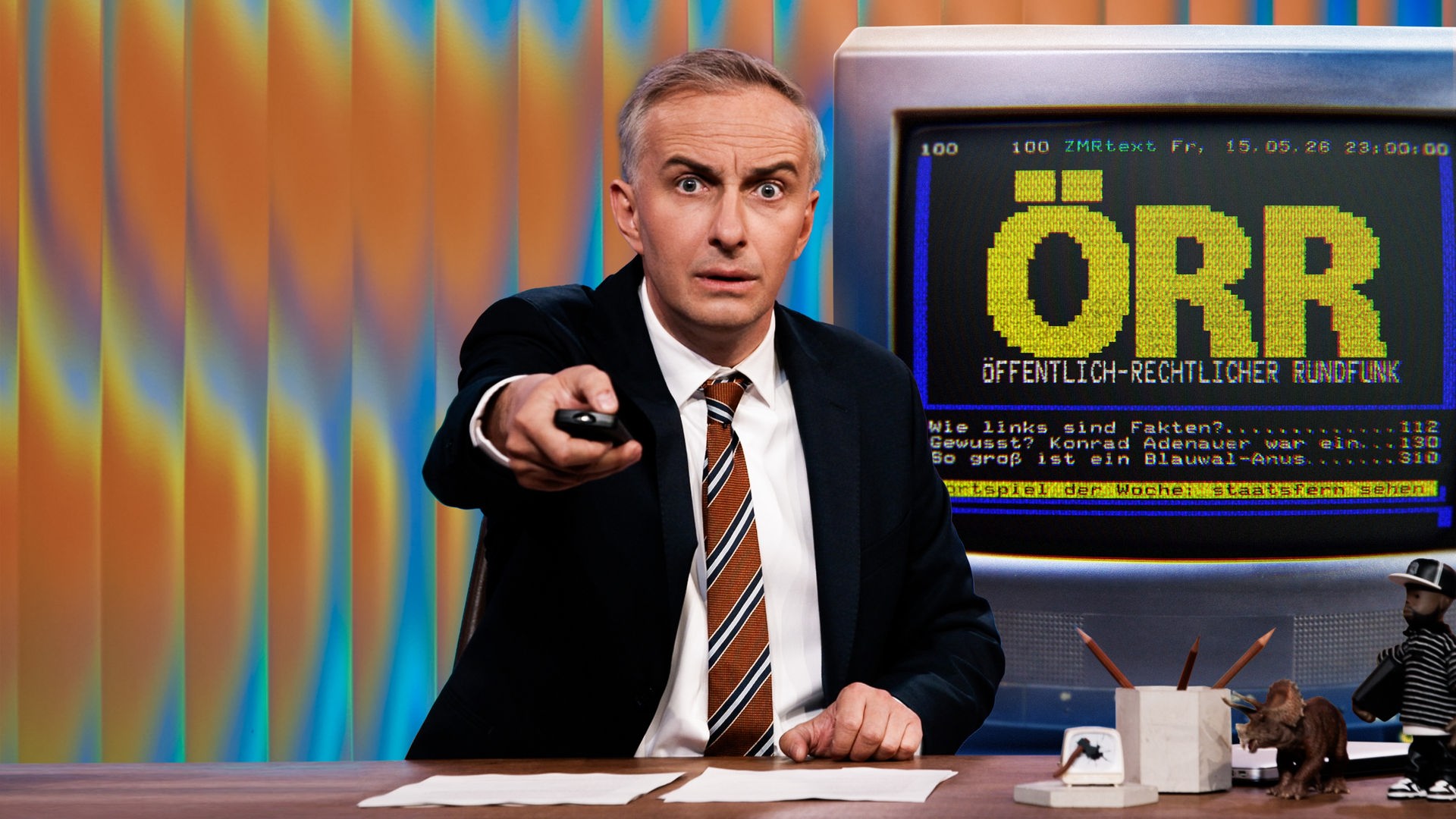 Jan Böhmermann sitzt am Schreibtisch und hält eine Fernbedienung in der Hand. Rechts im Hintergrund ist eine Grafik im Stil einer Videotext-Seite zu sehen. Dort steht in großen, gelben Buchstaben: "ÖRR" und in kleinerer Schrift darunter: "Öffentlich-rechtlicher Rundfunk". Im unteren Bereich sind drei Zeilen im Stil eines Videotext-Menüs zu sehen: "Wie links sind Fakten?", "Gewusst? Konrad Adenauer war ein…" und "So groß ist ein Blauwal-Anus".