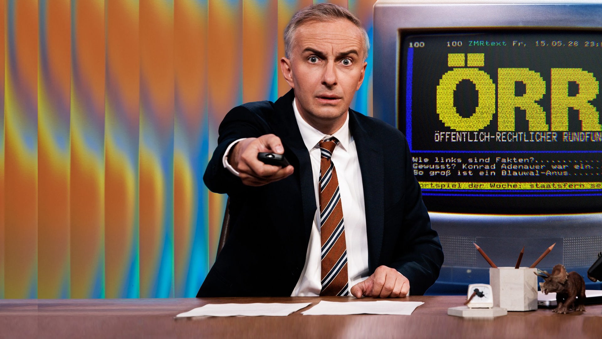 Jan Böhmermann sitzt am Schreibtisch und hält eine Fernbedienung in der Hand. Rechts im Hintergrund ist eine Grafik im Stil einer Videotext-Seite zu sehen. Dort steht in großen, gelben Buchstaben: "ÖRR" und in kleinerer Schrift darunter: "Öffentlich-rechtlicher Rundfunk". Im unteren Bereich sind drei Zeilen im Stil eines Videotext-Menüs zu sehen: "Wie links sind Fakten?", "Gewusst? Konrad Adenauer war ein…" und "So groß ist ein Blauwal-Anus".