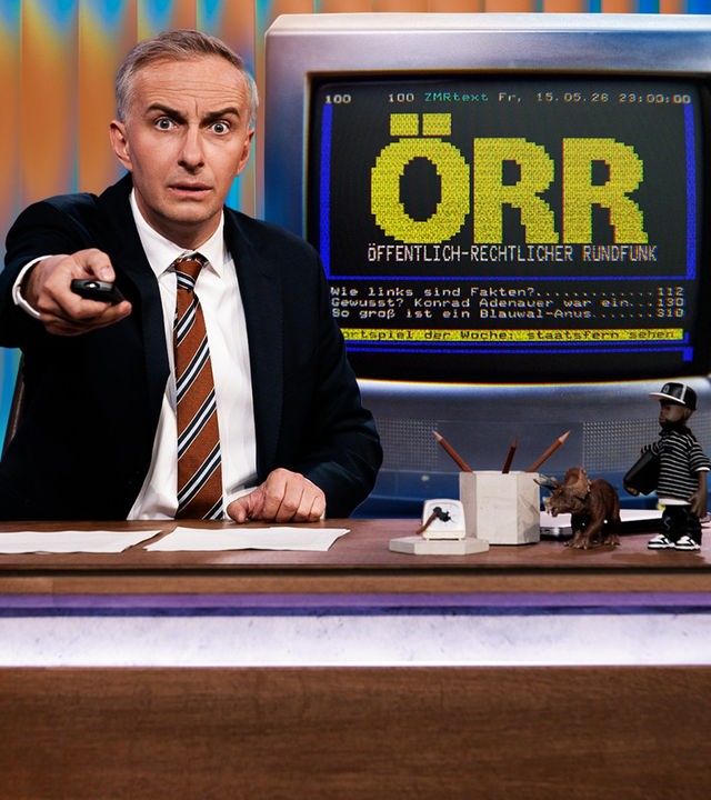 Jan Böhmermann sitzt am Schreibtisch und hält eine Fernbedienung in der Hand. Rechts im Hintergrund ist eine Grafik im Stil einer Videotext-Seite zu sehen. Dort steht in großen, gelben Buchstaben: "ÖRR" und in kleinerer Schrift darunter: "Öffentlich-rechtlicher Rundfunk". Im unteren Bereich sind drei Zeilen im Stil eines Videotext-Menüs zu sehen: "Wie links sind Fakten?", "Gewusst? Konrad Adenauer war ein…" und "So groß ist ein Blauwal-Anus".