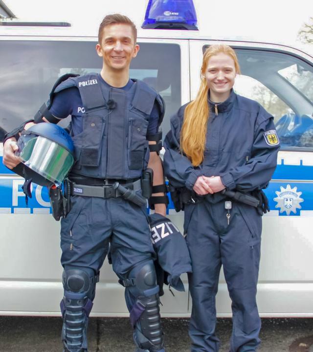 Eric steht in der Montur eines Polizisten vor einem Polizeiauto. Rechts neben ihm steht eine Polizstin.