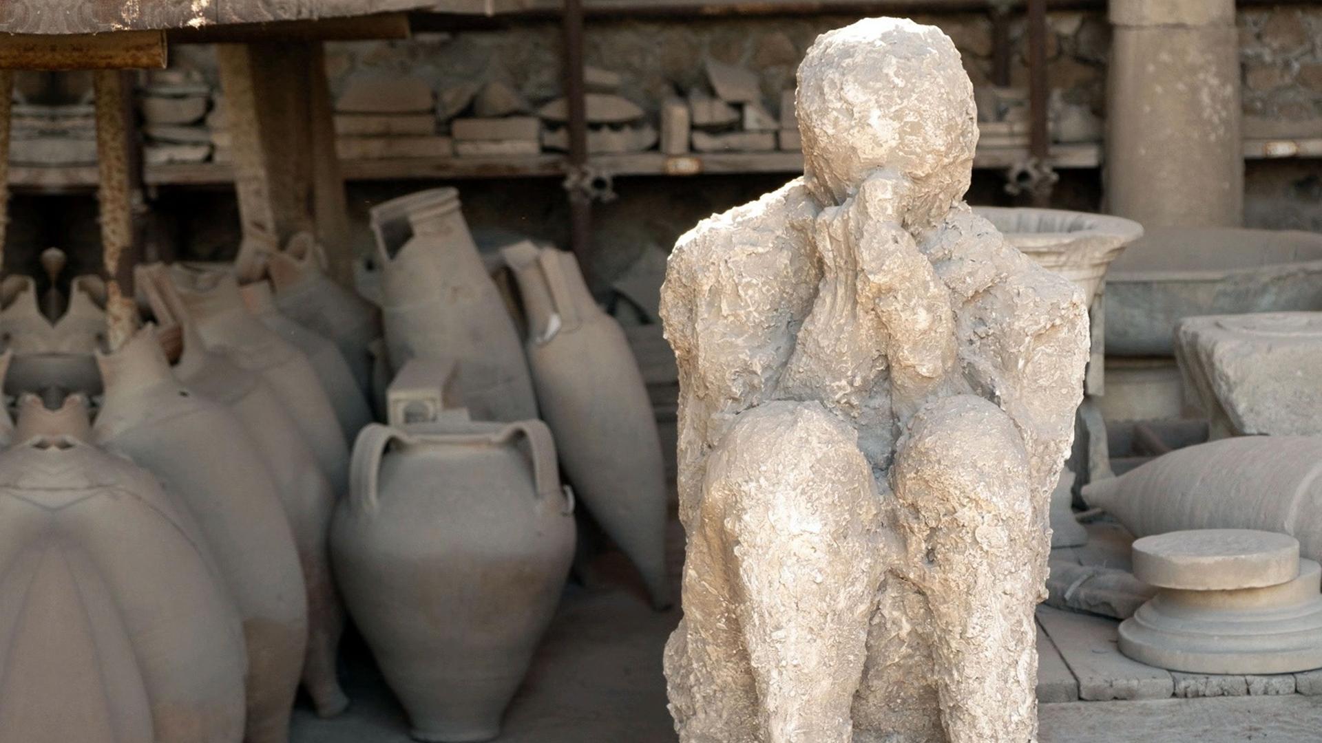 "The Pompeji Catastrophe": Von Tongefäßen umgebene, versteinerte menschliche Figur, die mit angezogenen Beinen auf dem Boden sitzt und die Hände zum Gebet vor das Gesicht hält.