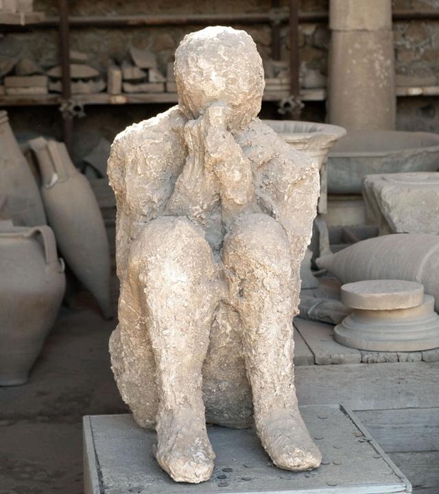 "The Pompeji Catastrophe": Von Tongefäßen umgebene, versteinerte menschliche Figur, die mit angezogenen Beinen auf dem Boden sitzt und die Hände zum Gebet vor das Gesicht hält.