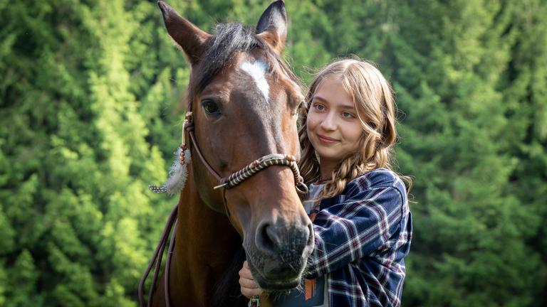 "Ponyherz - Wild und frei": Ponyherz und Anni stehen dicht beieinander. Ponyherz hat eine markante weiße Blesse auf der Stirn und trägt ein verziertes Halfter mit einer hellen Feder an der Seite. Anni steht leicht versetzt hinter dem Pferdekopf auf der rechten Seite. Sie blickt mit einem sanften Lächeln direkt zu Ponyherz. Im Hintergrund sind dichte, grüne Nadelbäume zu sehen.