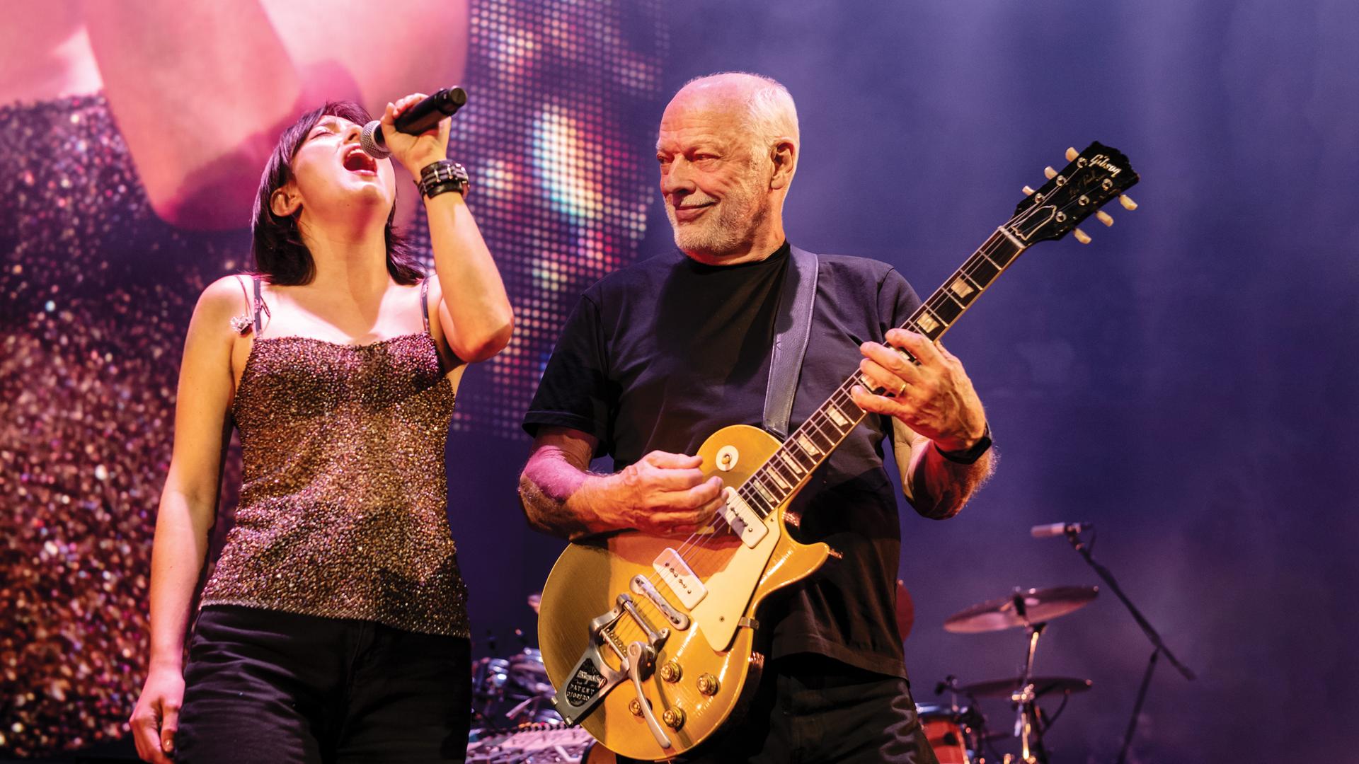 David Gilmour steht auf der Bühne und spielt Gitarre. Er blickt nach rechts zu einer Frau, die singt.