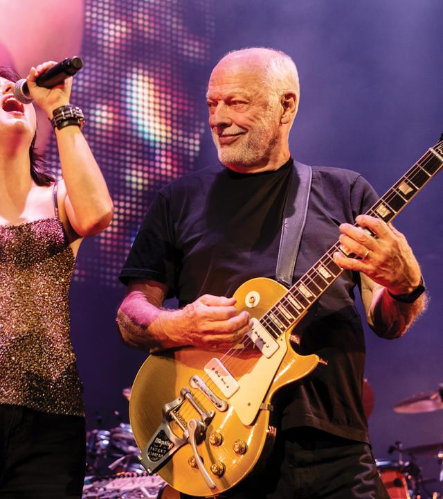 David Gilmour steht auf der Bühne und spielt Gitarre. Er blickt nach rechts zu einer Frau, die singt.