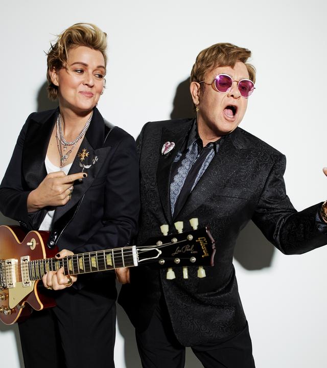 Brandi Carlile steht mit umgehängter Gitarre neben Elton John und schaut ihn an und zeigt auf ihn. Elton John trägt einen glitzernen Anzug und rosa Brille und singt.