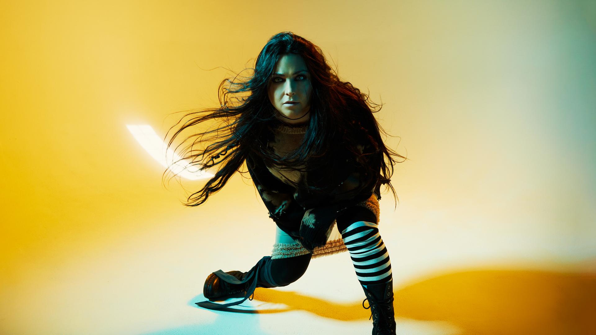 Vor einem gelben Hintergrund ist Amy Lee von Evanescence die mitten in einer schwungvollen Bewegung fotografiert wird. Ihre Haare fliegen durch die Luft und sie blickt in die Kamera