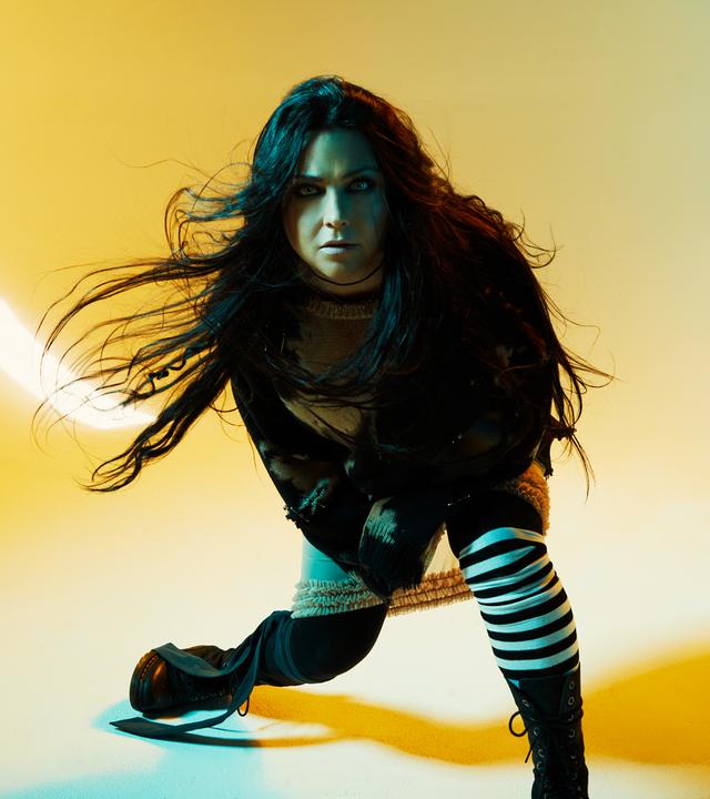 Vor einem gelben Hintergrund ist Amy Lee von Evanescence die mitten in einer schwungvollen Bewegung fotografiert wird. Ihre Haare fliegen durch die Luft und sie blickt in die Kamera