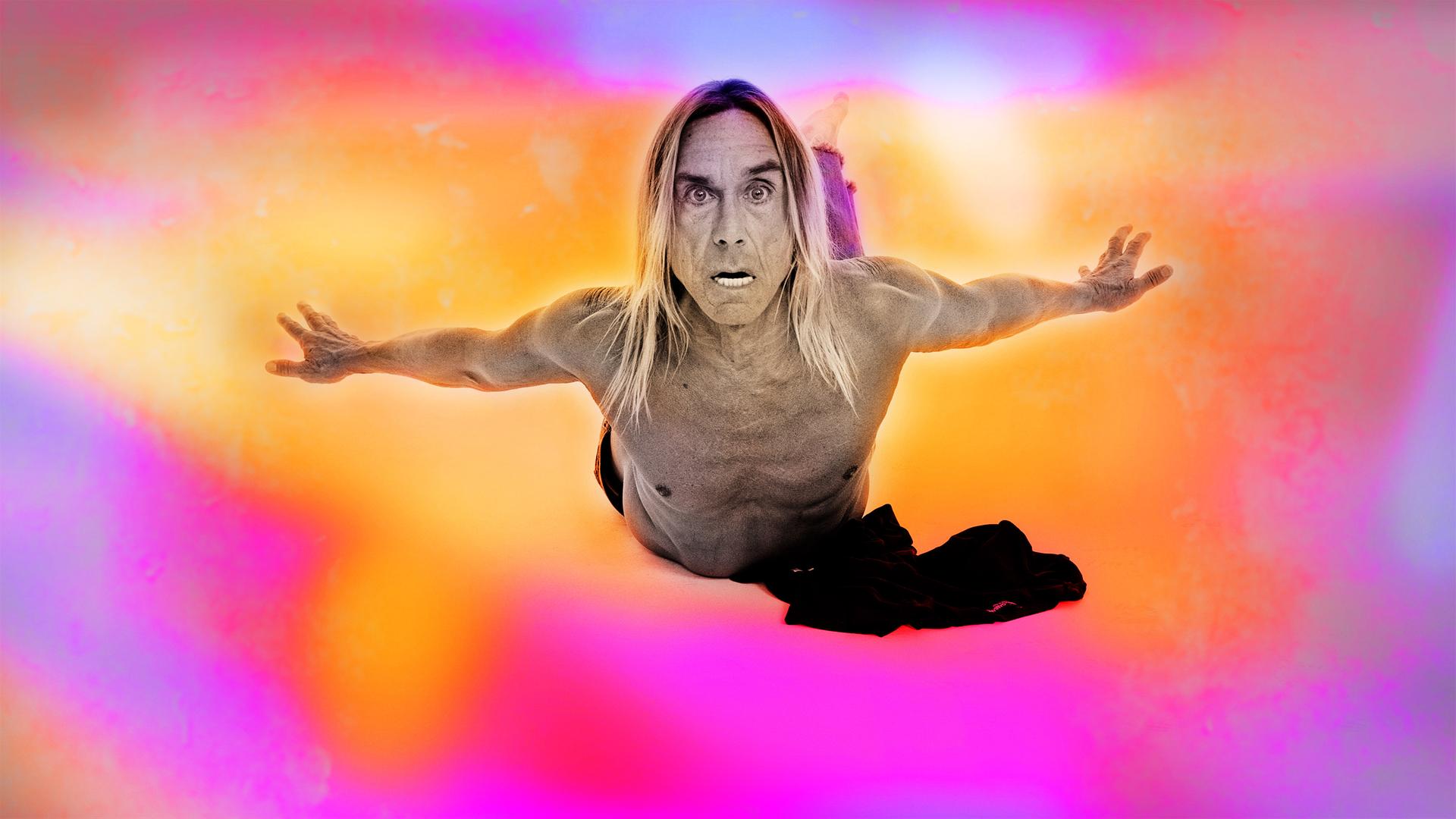 Iggy Pop fliegt oberkörperfrei mit Blick in die Kamera durch einen orange-pinken Himmel. 