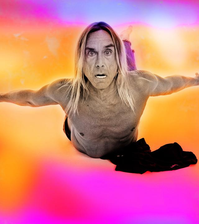 Iggy Pop fliegt oberkörperfrei mit Blick in die Kamera durch einen orange-pinken Himmel. 