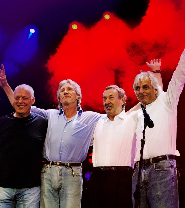 Vier Bandmitglieder von Pink Floyd stehen Arm in Arm auf einer Bühne.
