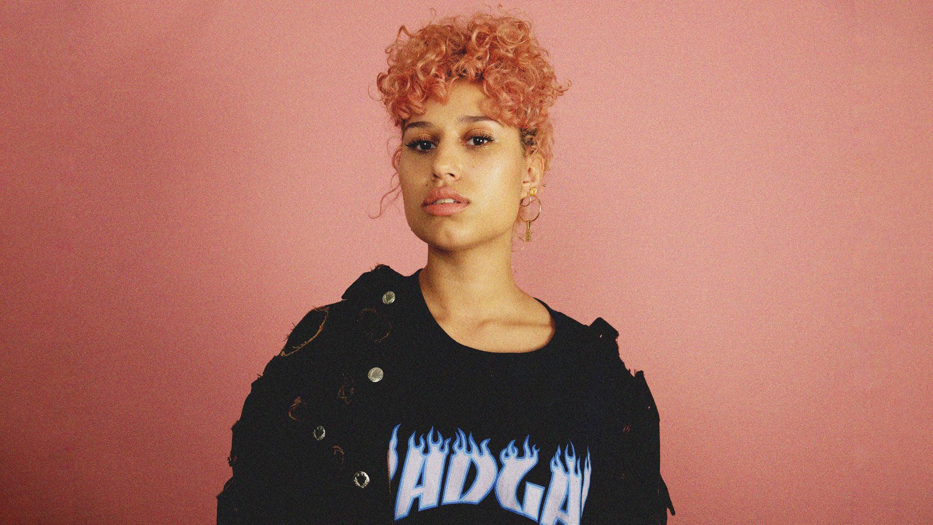 Raye steht mit rosa gelockten Haaren und schwarzem Shirt vor einer rosanen Wand und schaut in die Kamera.