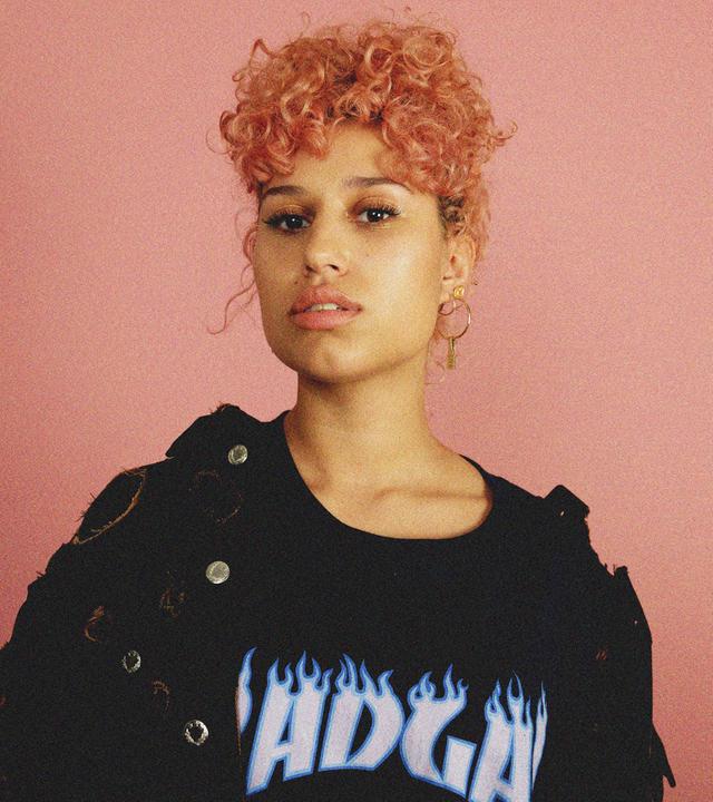 Raye steht mit rosa gelockten Haaren und schwarzem Shirt vor einer rosanen Wand und schaut in die Kamera.