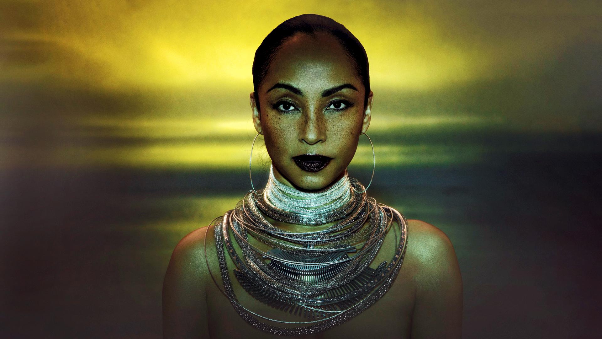 Sade Adu trägt große Kreolen und ganz viele Ketten um den Hals. Ihre Haare sind streng zurückgebunden und sie blickt in die Kamera.
