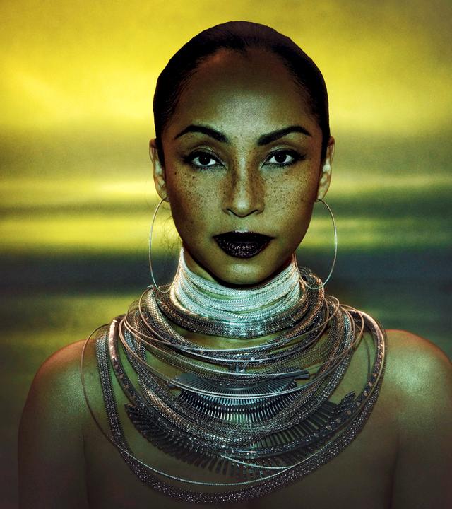 Sade Adu trägt große Kreolen und ganz viele Ketten um den Hals. Ihre Haare sind streng zurückgebunden und sie blickt in die Kamera.