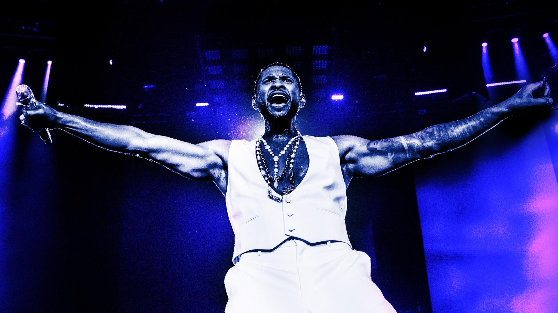 Usher steht mit offenen Armen und Mikro in der Hand auf der Bühne und singt.