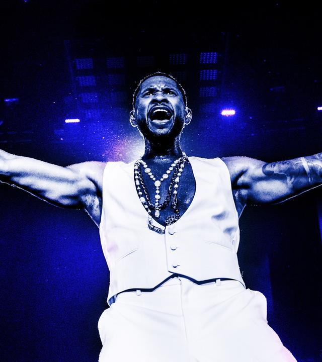 Usher steht mit offenen Armen und Mikro in der Hand auf der Bühne und singt.