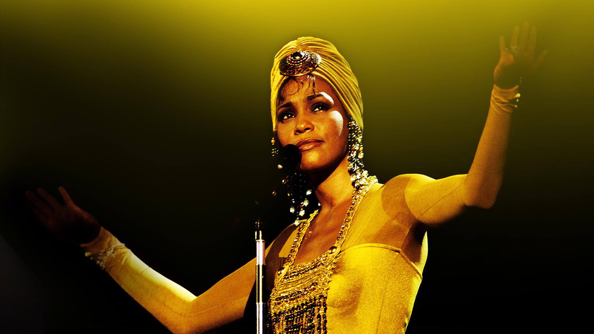 Whitney Houston steht vor einem Mikrofon, die Arme offen erhoben in der Luft. Die Haare trägt sie in einem Turban und sie trägt glitzernde Ohrringe und Oberteil.