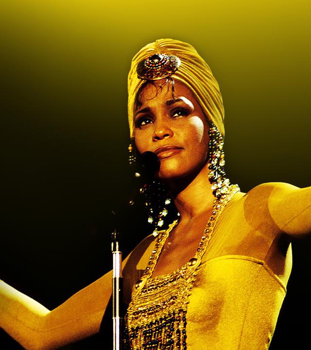 Whitney Houston steht vor einem Mikrofon, die Arme offen erhoben in der Luft. Die Haare trägt sie in einem Turban und sie trägt glitzernde Ohrringe und Oberteil.