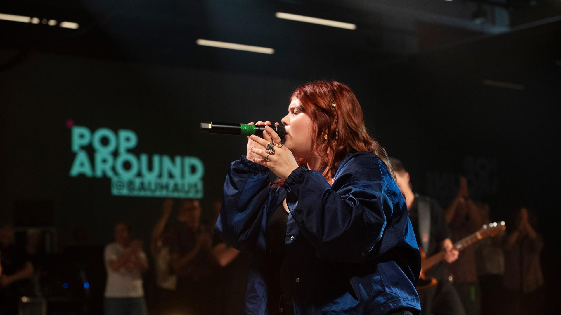 Loi singt ins Mirkofon. Hinter ist Publikum und ein der Pop Around Bauhaus Schriftzug zu sehen.