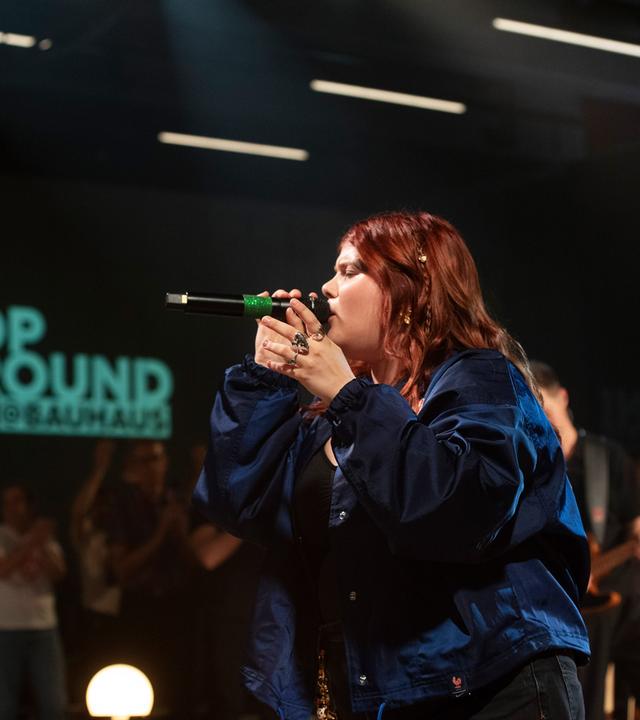 Loi singt ins Mirkofon. Hinter ist Publikum und ein der Pop Around Bauhaus Schriftzug zu sehen.