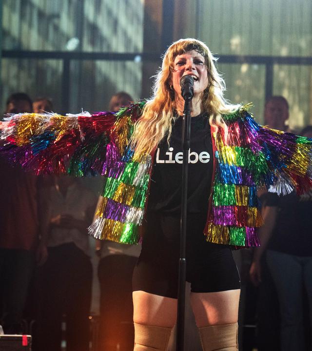 MIA steht mit offenen Armen und in regenbogenfarbener Glitzerfransenjacke und T-Shirt mit der Aufschrift "Liebe" auf der Bühne und singt ins Mikrofon. 