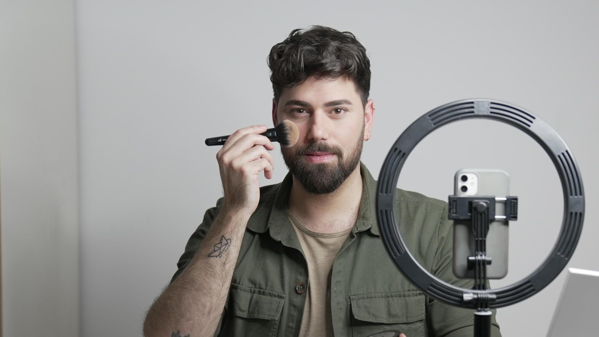 Davide (32) pudert sich die Wange, während er an einem Tisch sitzt, auf dem Schminkutensilien verteilt sind und filmt sich dabei mit dem Handy.