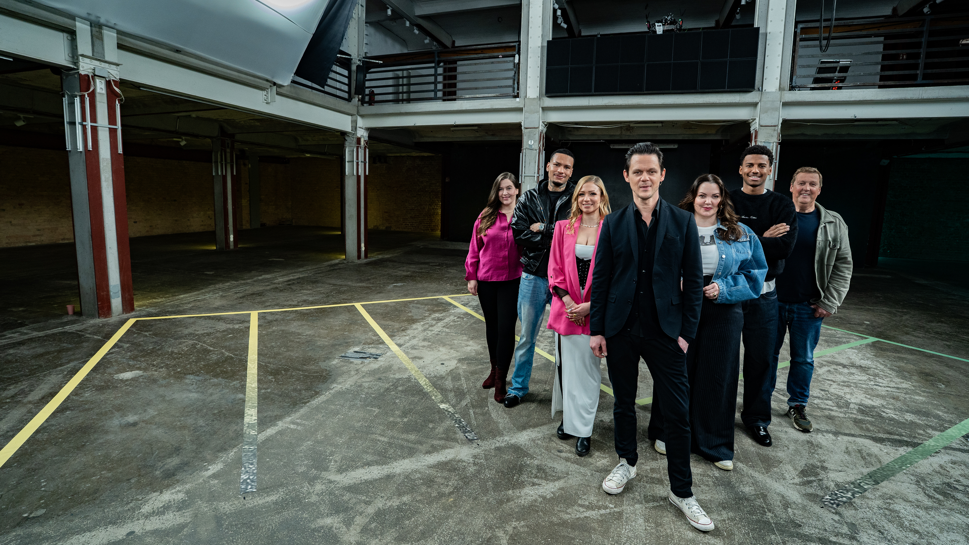 Es stehen sieben Menschen auf einem Boden, der ein schachbrettartiges Muster hat. Die sieben Menschen, drei Frauen und vier Männer, stehen in einer Reihe. Alle blicken in die Richtung des Betrachters.