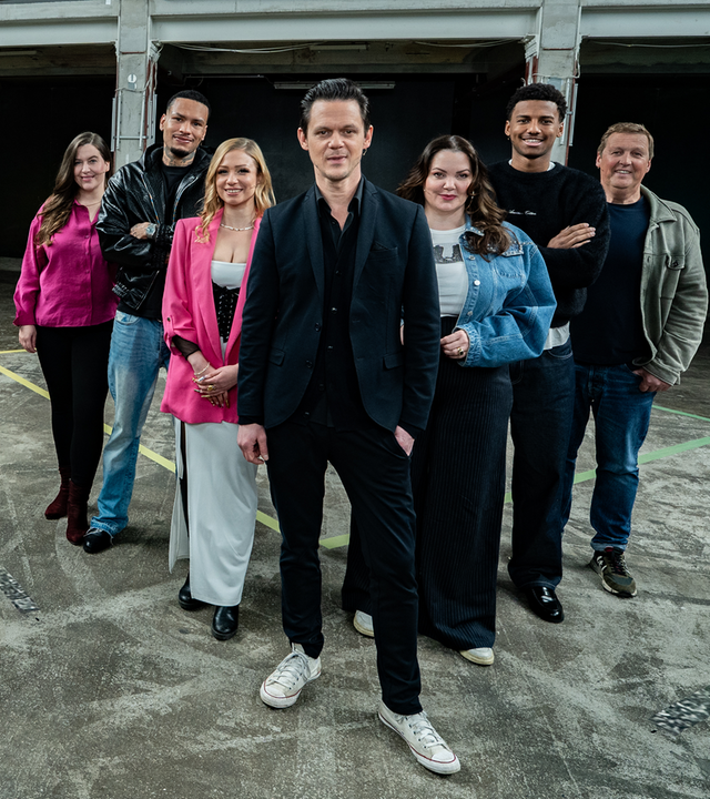 Es stehen sieben Menschen auf einem Boden, der ein schachbrettartiges Muster hat. Die sieben Menschen, drei Frauen und vier Männer, stehen in einer Reihe. Alle blicken in die Richtung des Betrachters.