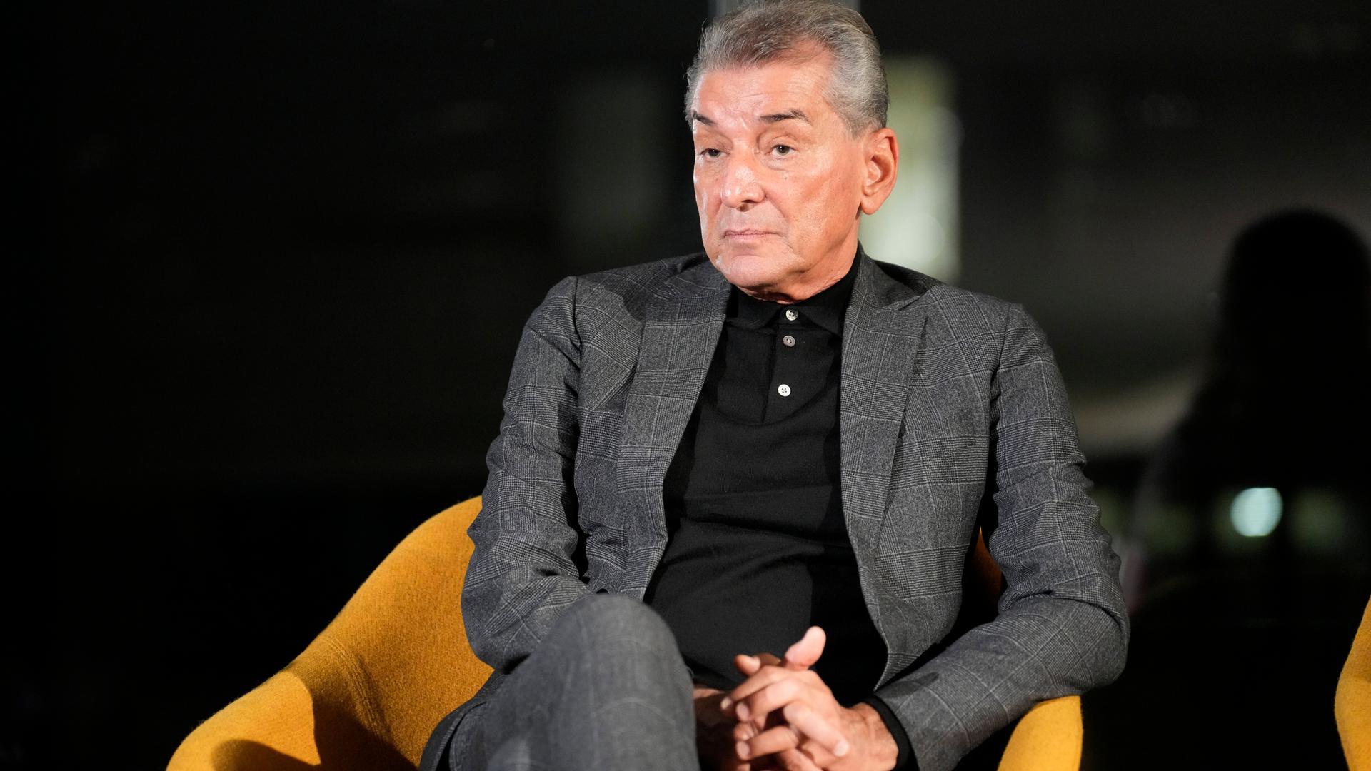 Ein älterer Mann im dunkelgrauen Anzug sitzt vor einem dunklen Hintergrund auf einem schwarzen Sessel.