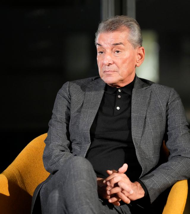 Ein älterer Mann im dunkelgrauen Anzug sitzt vor einem dunklen Hintergrund auf einem schwarzen Sessel.