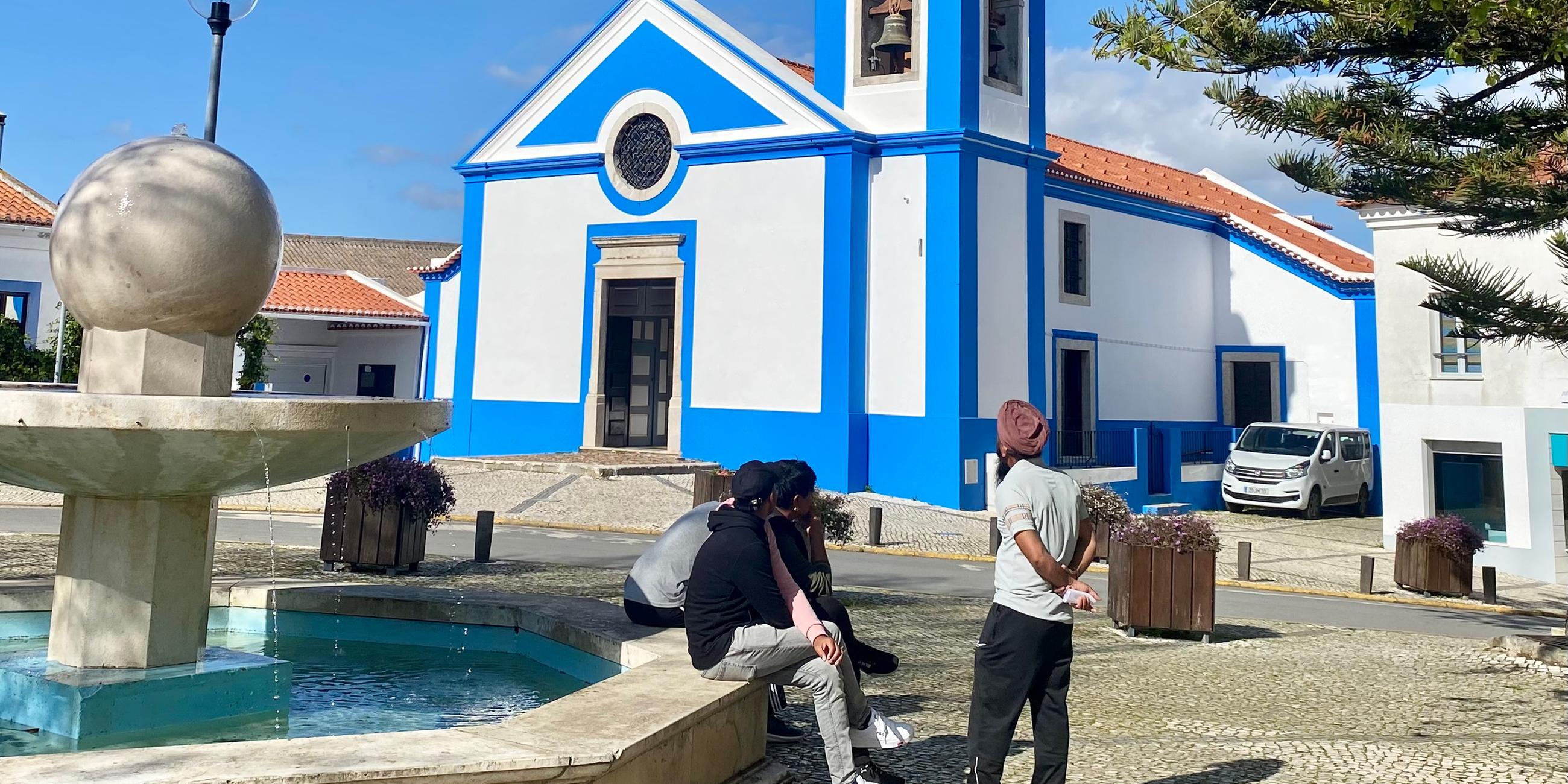 In einer portugisischen Stadt befindet sich auf dem Platz vor der Kirche ein Brunnen, an dessen Rand Menschen sitzen.