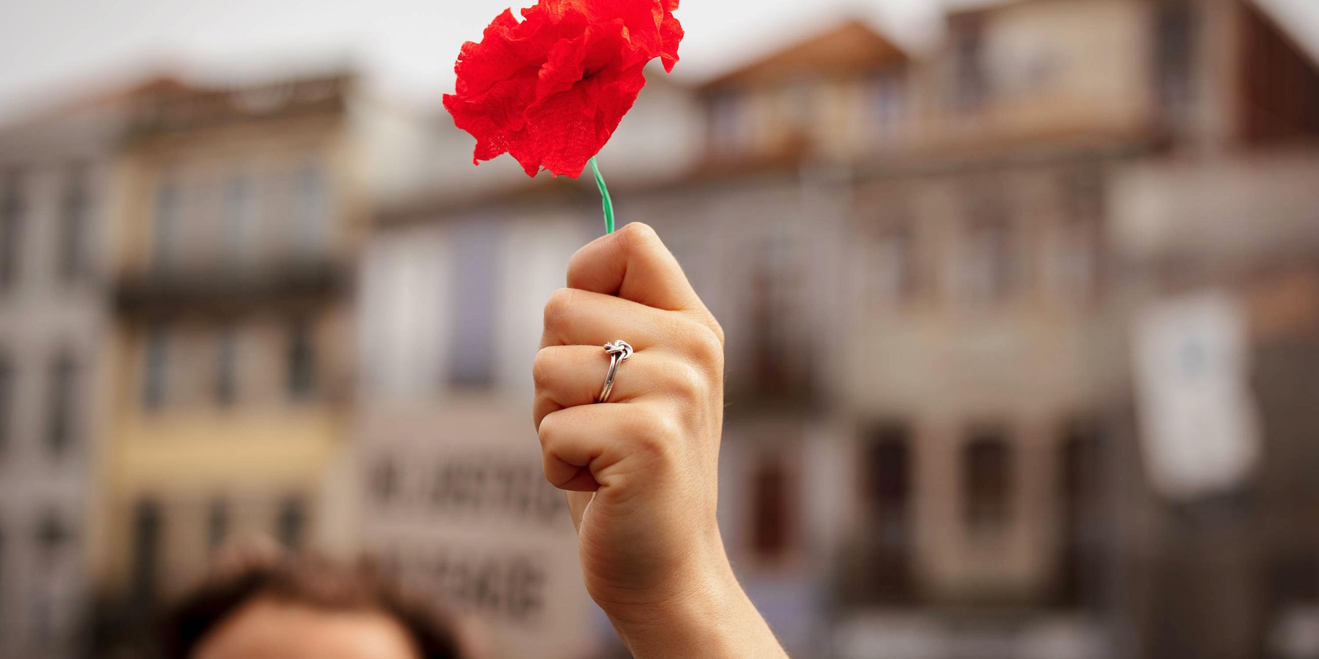 Eine Frauenhand mit einem Ring am Finger hält eine rote Nelke in die Höhe.